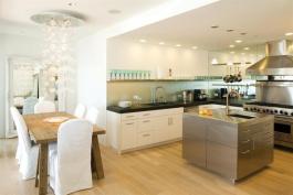 Cabinets are made from straightforward low-maintenance materials like MDF doors and drawer fronts finished with pre-catalyzed, integrated cream-colored lacquer. Elongated steel hardware matches appliance detailing. A dropped ceiling sets the kitchen alcove apart from the adjacent great room.