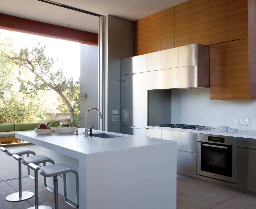 Stainless steel cabinets form a unified front with the stainless appliances. Teak cabinets above inject a note of warmth. The island picks up on the wall assembly's nested “L” shapes and Corian counter.