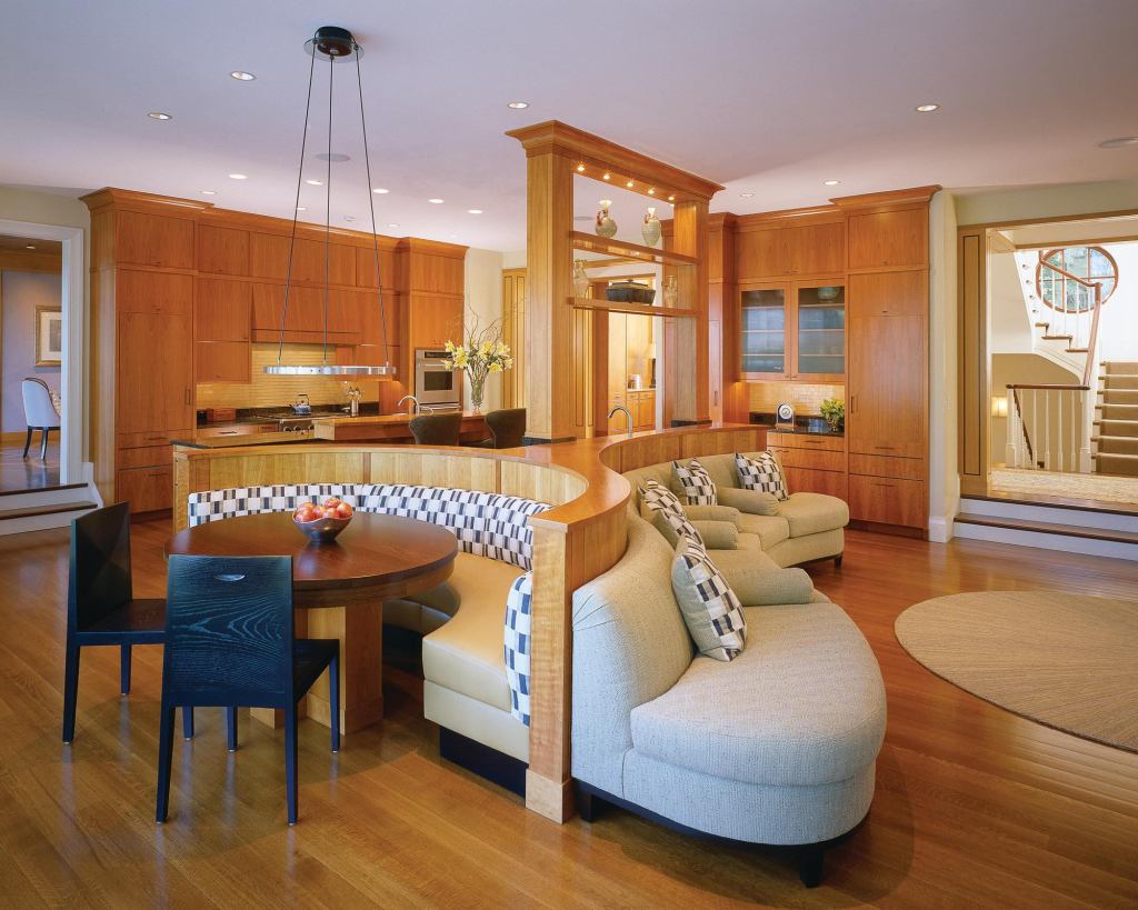 Anchoring a room that contains a kitchen, breakfast area, and family sitting area, the upholstered cherry piece turns a different face to each function: a counter with a bar sink toward the kitchen, a semicircular seating booth at the breakfast area, and a lower, compound-curved built-in sofa facing the family rooms fireplace.