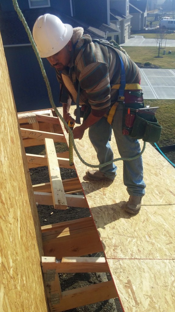 On a Summit Custom Homes jobsite, a framing carpenter wears a safety harness secured to a point above him while working on the roof. He is wearing a hardhat and shoes that have good traction for the slope of the
roof.  