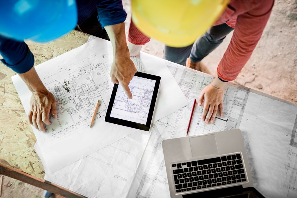 Female architect discussing construction plans with foreman on paper, digital tablet and laptop. Overhead shot.