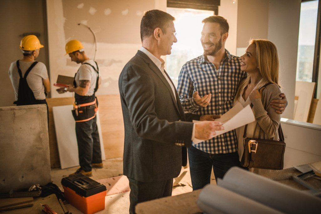 Young happy couple talking to their investor about housing plan inside of an apartment. Workers are in the background.