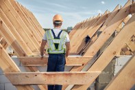 Back-view photo of a man in a hardhat and high-visibility vest standing near a wooden roof frame