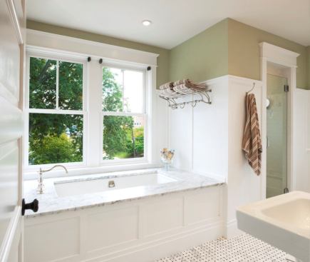 The bathtub is on a south wall, taking advantage of sunlight. Double-hung windows are thermally paned and energy efficient. The bath isn't large, yet it includes a two-headed shower, tub, twin pedestal sinks, a toilet, and a free-standing vanity. Three-quarter height wainscoting and a darker paint color on the upper wall make the space strong and intimate.