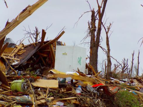 A Twister Safe tornado shelter sits attached to its concrete base slab amid the remnants of a completely destroyed house in Joplin after the tornado. Shattered trees and clean-swept house lots indicate an F-5 degree of tornado damage; the Twister Safe is unscathed. The winter following the tornado, Twister Safe’s owners celebrated with more than two dozen customers whose lives were protected by the units during the storm.