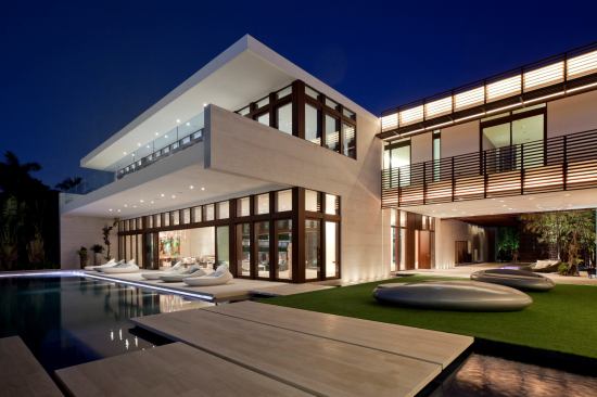 Seen from the rear, the home features laminated mahogany windows and door frames, water elements, and a glass-fronted second level. The metal brise-soleil, canopies, and louvered walls feature a powder-coated paint in a woodgrain texture.