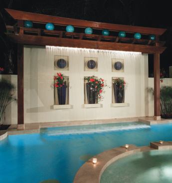 The privacy wall is pushed to the lot’s setback line to create as much space for the courtyard as possible.The wall’s centerpiece is topped with a trellis that hides the apparatus for a rain curtain; the waterfall box is sandwiched between two small beams that appear to be a single large one. Fiber-optic lighting adds drama to the falling water and spotlights the colorful glazed ceramic planters and spheres in the wall’s niches.