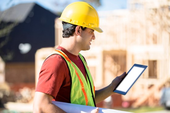 A Latin descent construction worker, supervisor, contractor, inspector, architect, or engineer inspects the work at a construction site. Framed house, building in background. He is wearing a hard hat and safety vest and holds a digital tablet and blueprints.