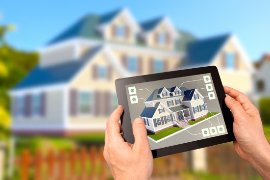 Choosing a new house to buy: hands holding a tablet showing a real estate listing with model of a new country home to buy, with labels pointing at features, and the potential house deal on the background. Buying dream house, real estate business and investment, home automation.