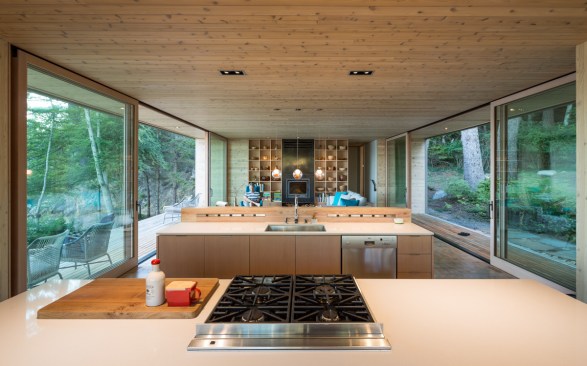 The open-plan kitchen and living and dining rooms are lined in wood paneling and custom, built-in shelves that connect the interior to the forest outside.