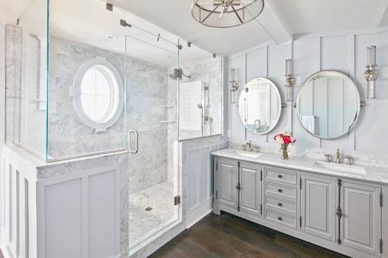 A large, glass-enclosed shower with marble tile and oval window stands as the focal point of the luxurious master bath.