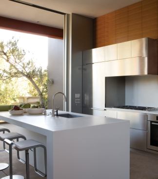 Steel cabinets form a unified front with the stainless appliances. Teak cabinets above inject a note of warmth. The island picks up on the wall assembly's nested L shapes and Corian counter.

