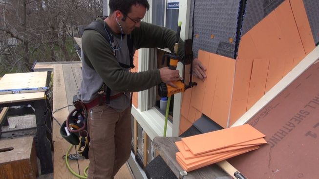 A carpenter staples pre-primed cedar shingles over a woven air-gap mesh.