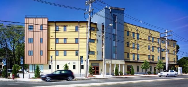 Red horizontal trim on the ochre fiber-cement siding reflects the highway’s streaming aesthetic. Copper-panel bays wrap the corners.