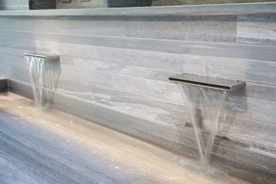 Above the wide trough sink and embedded in the stone wall are twin waterfall faucets.